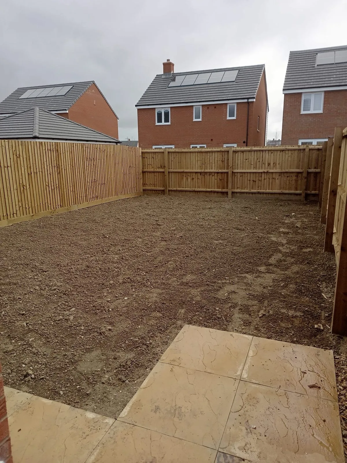 A tired lawn with long grass and a brown patch, seen from a patio toward a timber fence